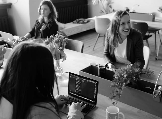 Three women sharing the same workspace, one is focused on some task, one is smiling at someone and one is typing something